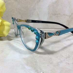 May include: Clear-framed eyeglasses with turquoise accents. The frames have a clear front and turquoise arms, embellished with turquoise-colored stones. The temples are black with gold-colored hinges. The glasses are labeled "R622 CHINA".