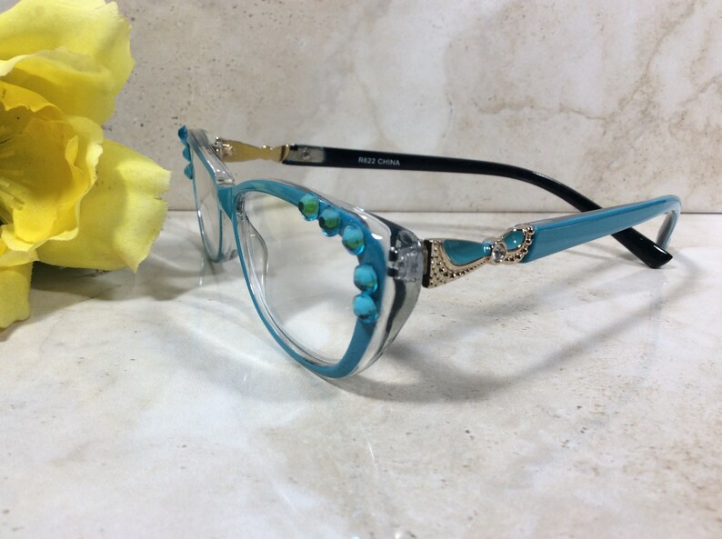 May include: Clear-framed eyeglasses with turquoise accents. The frames have a clear front and turquoise arms, embellished with turquoise-colored stones. The temples are black with gold-colored hinges. The glasses are labeled "R622 CHINA".