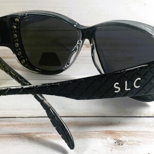 May include: Black sunglasses with dark lenses. The frames are glossy black with a quilted pattern on the arms. The text "SLC" is visible on the arm. The sunglasses have a decorative stud detail on the frame.