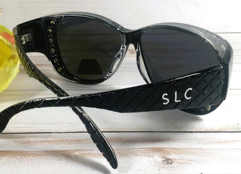 May include: Black sunglasses with dark lenses. The frames are glossy black with a quilted pattern on the arms. The text "SLC" is visible on the arm. The sunglasses have a decorative stud detail on the frame.