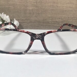 May include: Rectangular eyeglasses with a patterned black, red, and white frame. The frame is accented with gold-colored studs on the outer corners. The lenses are clear, and the glasses are set against a neutral background.