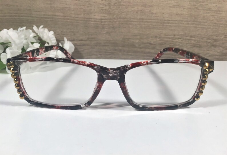 May include: Rectangular eyeglasses with a patterned black, red, and white frame. The frame is accented with gold-colored studs on the outer corners. The lenses are clear, and the glasses are set against a neutral background.