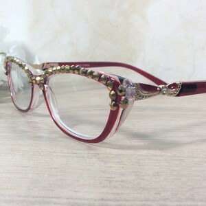 May include: Cat-eye style eyeglasses with red and clear frames, adorned with gold-coloured decorative elements. The top rim and temples feature the embellishments. The temples are a deep red, and the glasses are set against a light background.