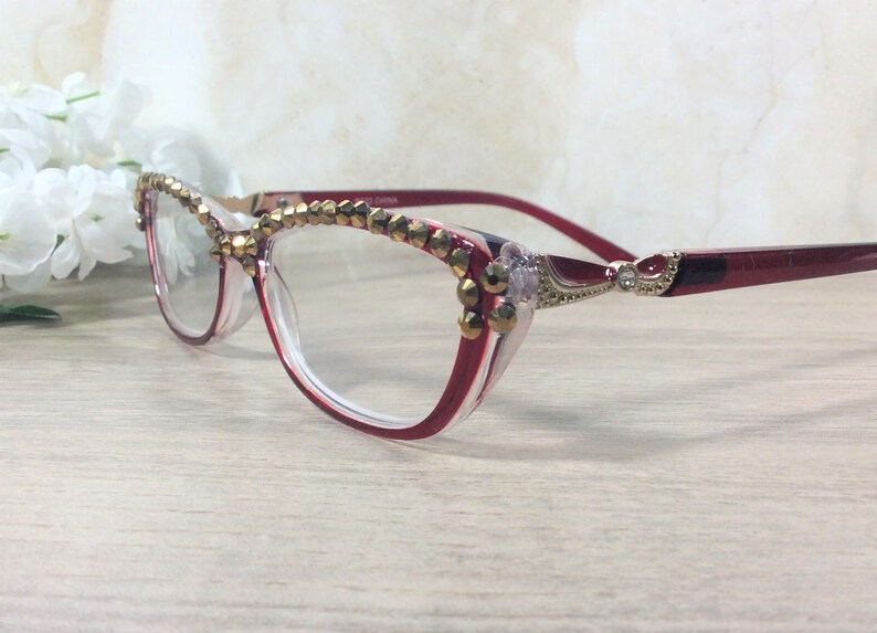 May include: Cat-eye style eyeglasses with red and clear frames, adorned with gold-coloured decorative elements. The top rim and temples feature the embellishments. The temples are a deep red, and the glasses are set against a light background.