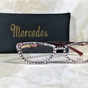 May include: Red-framed eyeglasses with clear lenses, embellished with sparkling rhinestones along the top and bottom rims. A black case with the gold text "Mercedes" is behind the glasses. The glasses have a +1.75 magnification.