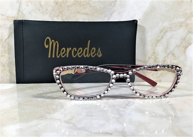 May include: Red-framed eyeglasses with clear lenses, embellished with sparkling rhinestones along the top and bottom rims. A black case with the gold text "Mercedes" is behind the glasses. The glasses have a +1.75 magnification.