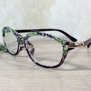 May include: Eyeglasses with a patterned frame, adorned with green and blue rhinestones along the top rim. The glasses have a gold-colored accent on the side and black arms. The lenses are clear, and the frame has a brown and white pattern.