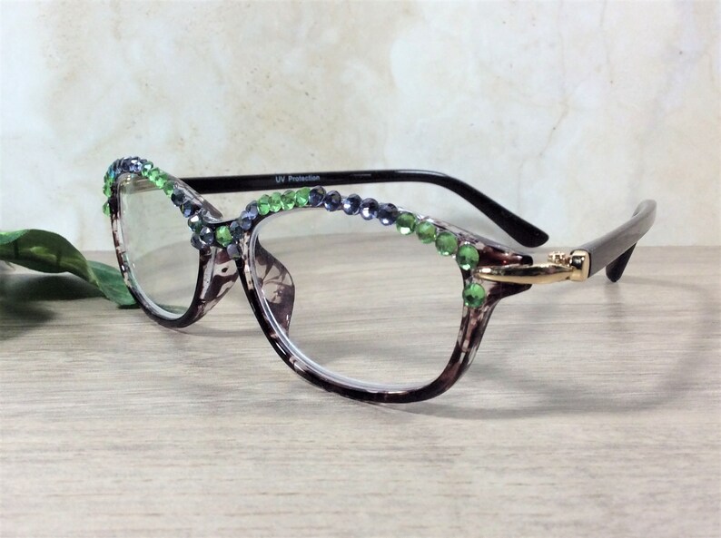 May include: Eyeglasses with a patterned frame, adorned with green and blue rhinestones along the top rim. The glasses have a gold-colored accent on the side and black arms. The lenses are clear, and the frame has a brown and white pattern.