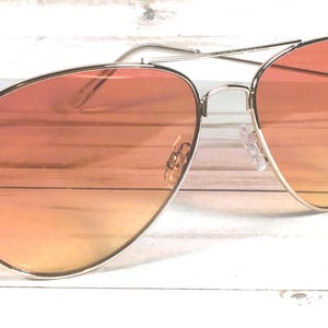 May include: Aviator sunglasses with a silver frame and orange gradient lenses. The lenses transition from a darker orange at the top to a lighter orange at the bottom. The sunglasses are set against a white wooden background.