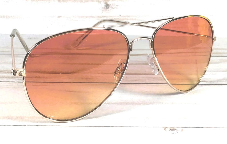 May include: Aviator sunglasses with a silver frame and orange gradient lenses. The lenses transition from a darker orange at the top to a lighter orange at the bottom. The sunglasses are set against a white wooden background.