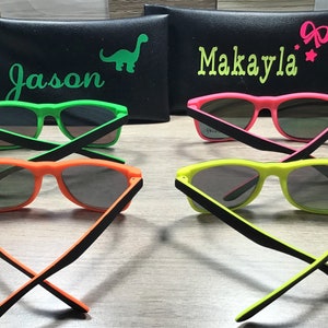 May include: Four pairs of neon-coloured sunglasses with black frames. The sunglasses are arranged in two pairs, with the arms crossed. The sunglasses are on a wooden surface. The sunglasses are labelled with the names "Jason" and "Makayla".