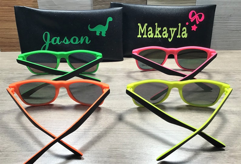 May include: Four pairs of neon-coloured sunglasses with black frames. The sunglasses are arranged in two pairs, with the arms crossed. The sunglasses are on a wooden surface. The sunglasses are labelled with the names "Jason" and "Makayla".