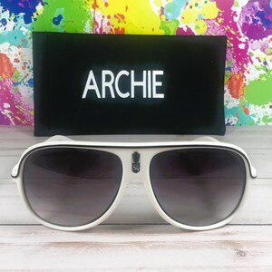 Puede incluir: Gafas de sol de aviador blancas con lentes oscuras y ribete negro. Las gafas de sol tienen un peque&ntilde;o emblema en la montura y se combinan con una funda negra que dice "ARCHIE" en letras blancas. El fondo presenta un dise&ntilde;o de salpicaduras colorido.