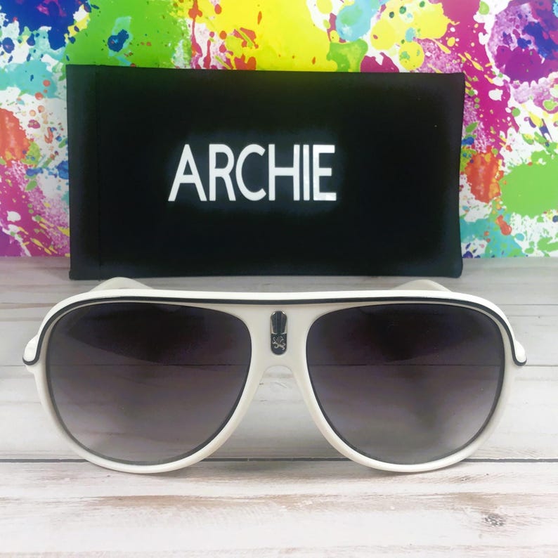 Puede incluir: Gafas de sol de aviador blancas con lentes oscuras y ribete negro. Las gafas de sol tienen un peque&ntilde;o emblema en la montura y se combinan con una funda negra que dice "ARCHIE" en letras blancas. El fondo presenta un dise&ntilde;o de salpicaduras colorido.