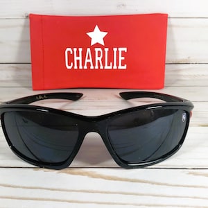 Op de afbeelding: Zwarte zonnebril met rode accenten en een sterlogo aan de zijkant. De zonnebril wordt geleverd met een rode etui met "CHARLIE" in witte letters.