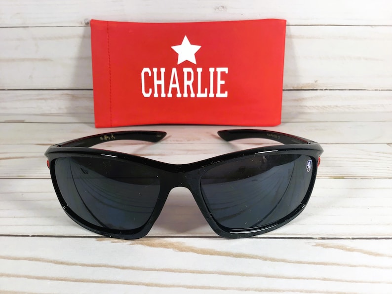 Op de afbeelding: Zwarte zonnebril met rode accenten en een sterlogo aan de zijkant. De zonnebril wordt geleverd met een rode etui met "CHARLIE" in witte letters.