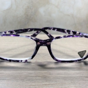 K&ouml;nnte beinhalten: Lesebrille mit rechteckigem Rahmen und lila-wei&szlig;em Muster. Die Brille hat kleine Strasssteine an den Ecken und eine Vergr&ouml;&szlig;erung von +3,00. Die Brille liegt auf einer Holzoberfl&auml;che.