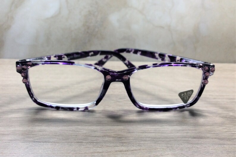 K&ouml;nnte beinhalten: Lesebrille mit rechteckigem Rahmen und lila-wei&szlig;em Muster. Die Brille hat kleine Strasssteine an den Ecken und eine Vergr&ouml;&szlig;erung von +3,00. Die Brille liegt auf einer Holzoberfl&auml;che.