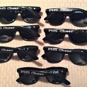 May include: Black sunglasses with the text "PHS Cheer" printed in white on the temples. Some sunglasses have names and stars printed on the temples. The sunglasses are a promotional item.