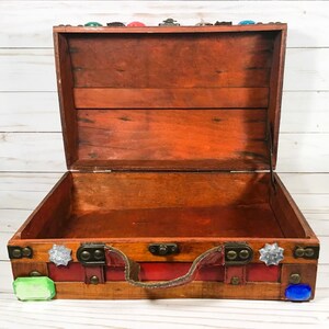 May include: Open wooden chest with a reddish-brown finish, brass hardware, and a red leather handle. The chest is decorated with colorful gemstones and has a hinged lid. The interior is empty.