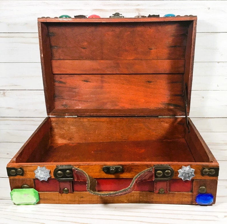 May include: Open wooden chest with a reddish-brown finish, brass hardware, and a red leather handle. The chest is decorated with colorful gemstones and has a hinged lid. The interior is empty.