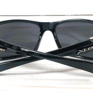 May include: Black sunglasses with dark lenses and a glossy finish. The temples are crossed, revealing the "XLOOP" logo in white. Silver accents adorn the hinges. The sunglasses are designed for outdoor activities and offer UV protection.