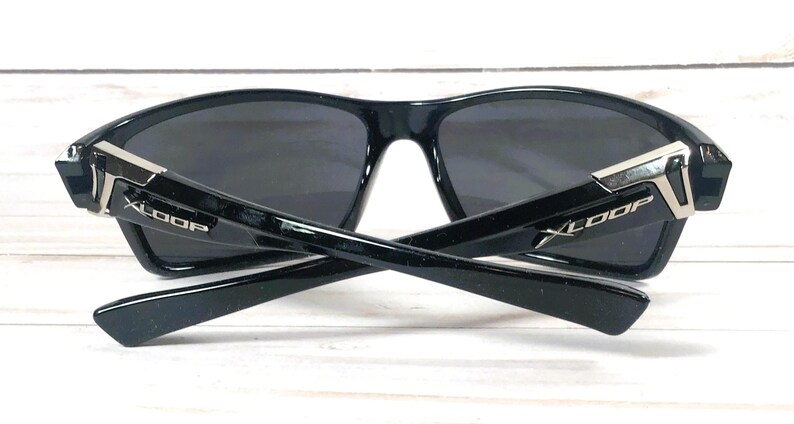 May include: Black sunglasses with dark lenses and a glossy finish. The temples are crossed, revealing the "XLOOP" logo in white. Silver accents adorn the hinges. The sunglasses are designed for outdoor activities and offer UV protection.