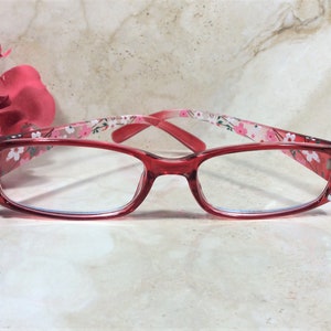 Puede incluir: Gafas con montura roja y lentes transparentes. Las patillas presentan un estampado floral y est&aacute;n adornadas con pedrer&iacute;a. Las gafas est&aacute;n sobre un fondo de m&aacute;rmol, con una flor rosa en la esquina superior izquierda.