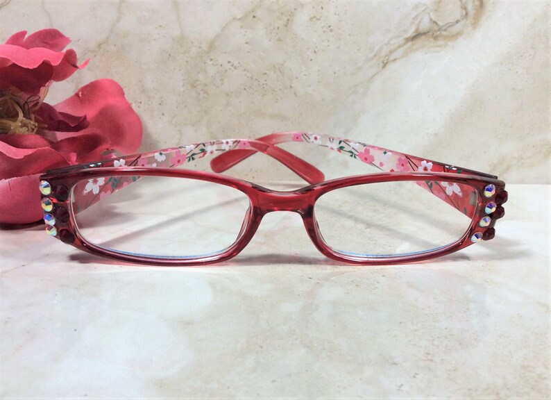Puede incluir: Gafas con montura roja y lentes transparentes. Las patillas presentan un estampado floral y est&aacute;n adornadas con pedrer&iacute;a. Las gafas est&aacute;n sobre un fondo de m&aacute;rmol, con una flor rosa en la esquina superior izquierda.