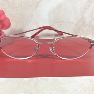 Pu&ograve; includere: Occhiali da lettura con montatura argentata, lenti e aste rosse. Le lenti ovali sono decorate con piccoli gioielli rossi sui bordi esterni. Gli occhiali hanno un ingrandimento di +2,25.