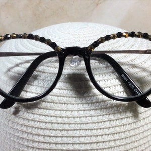 May include: Round, black-framed spectacles with clear lenses. The upper rims are decorated with small, dark beads. The glasses feature a metal bridge and the text "R664 +225 CHINA" is printed on the arm.