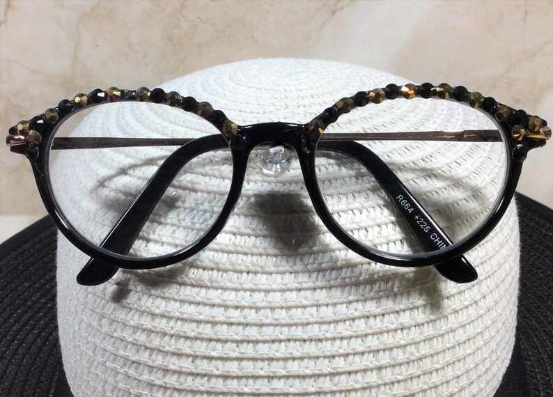 May include: Round, black-framed spectacles with clear lenses. The upper rims are decorated with small, dark beads. The glasses feature a metal bridge and the text "R664 +225 CHINA" is printed on the arm.