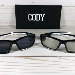 May include: Two pairs of black sunglasses with white accents. The sunglasses have a rectangular shape with a mirrored lens. The sunglasses are on a white wooden surface. The sunglasses are in a black case with the name "Cody" printed on it.