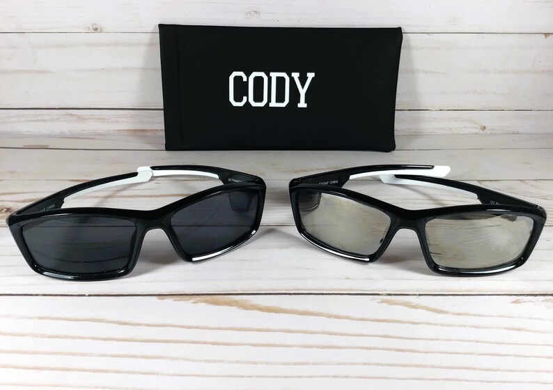 May include: Two pairs of black sunglasses with white accents. The sunglasses have a rectangular shape with a mirrored lens. The sunglasses are on a white wooden surface. The sunglasses are in a black case with the name "Cody" printed on it.