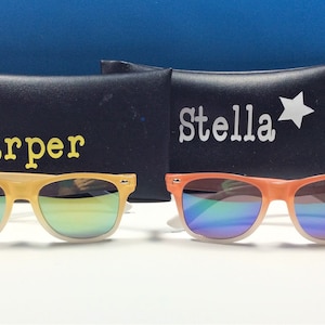 May include: Two pairs of sunglasses with mirrored lenses and colorful frames. The sunglasses are in their cases, which are black with white text. The case on the left says "Harper" and the case on the right says "Stella" with a star.