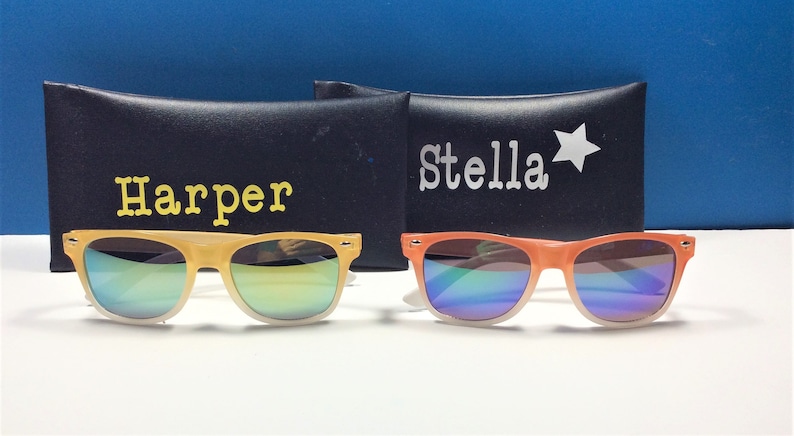 May include: Two pairs of sunglasses with mirrored lenses and colorful frames. The sunglasses are in their cases, which are black with white text. The case on the left says "Harper" and the case on the right says "Stella" with a star.