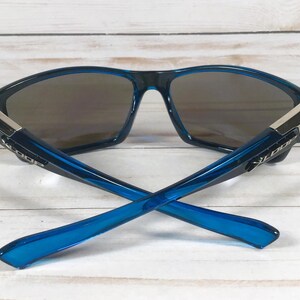 May include: Blue sunglasses with a dark lens and a modern design. The frames are a vibrant blue with silver accents at the temples. The arms of the sunglasses are crossed, showcasing the sleek design.