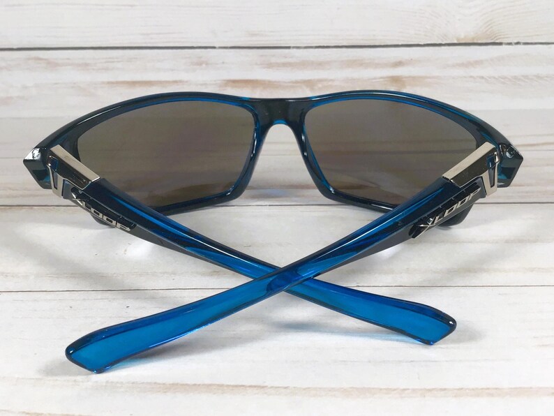 May include: Blue sunglasses with a dark lens and a modern design. The frames are a vibrant blue with silver accents at the temples. The arms of the sunglasses are crossed, showcasing the sleek design.