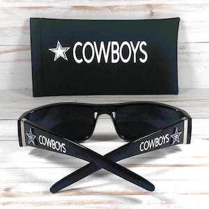 May include: Black sunglasses with the word "COWBOYS" and a star logo on the temples. A black case with the word "COWBOYS" and a star logo is behind the sunglasses. The sunglasses and case are on a light wood surface.
