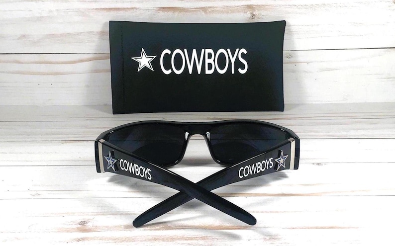 May include: Black sunglasses with the word "COWBOYS" and a star logo on the temples. A black case with the word "COWBOYS" and a star logo is behind the sunglasses. The sunglasses and case are on a light wood surface.