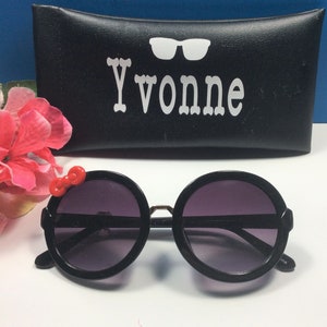 May include: Black round sunglasses with a red bow detail on the side. The sunglasses are in a black case with the name "Yvonne" printed on it.