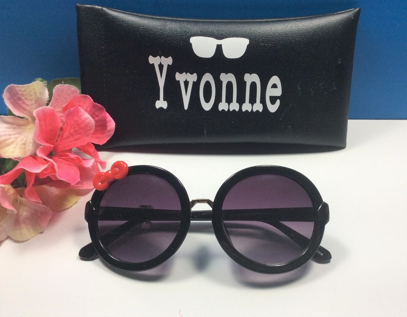 May include: Black round sunglasses with a red bow detail on the side. The sunglasses are in a black case with the name "Yvonne" printed on it.