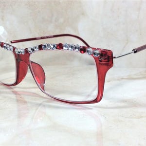 May include: Red-framed eyeglasses with clear lenses. The top of the frame is embellished with a row of clear and red rhinestones. The temples are a darker red color. The glasses are set against a neutral background.