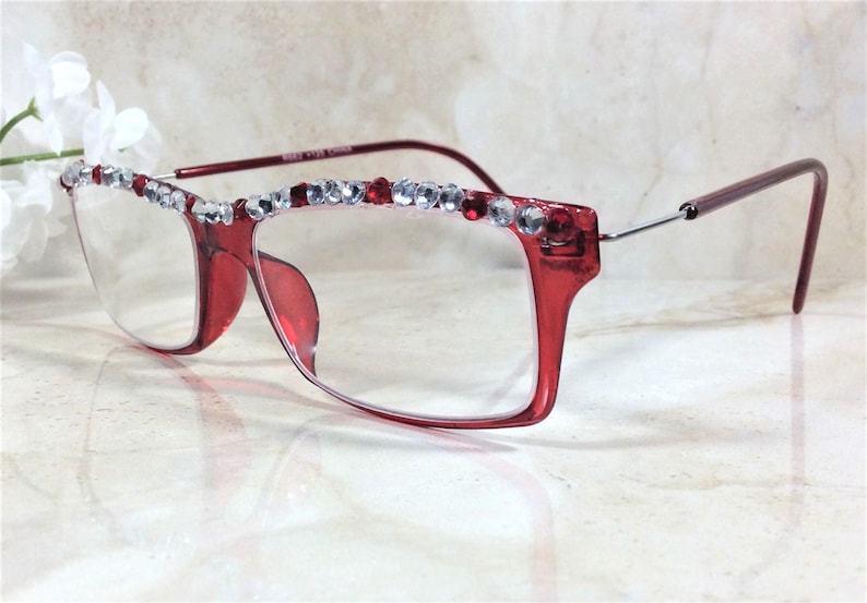 May include: Red-framed eyeglasses with clear lenses. The top of the frame is embellished with a row of clear and red rhinestones. The temples are a darker red color. The glasses are set against a neutral background.