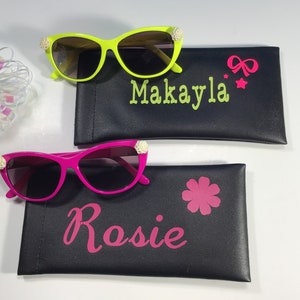 May include: Two pairs of sunglasses with decorative embellishments, each in a different colour, with personalised cases. The green sunglasses have a case with the name "Makayla" and a pink bow and stars. The pink sunglasses have a case with the name "Rosie" and a pink flower.