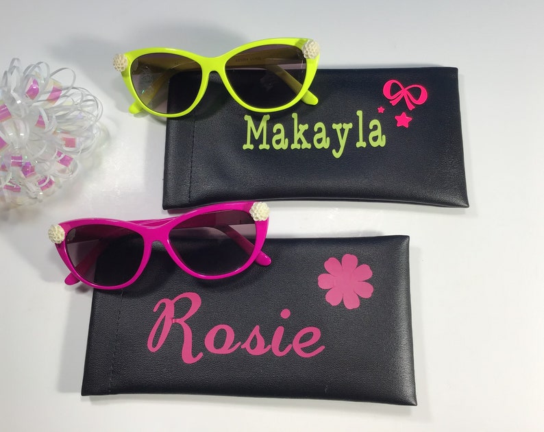 May include: Two pairs of sunglasses with decorative embellishments, each in a different colour, with personalised cases. The green sunglasses have a case with the name "Makayla" and a pink bow and stars. The pink sunglasses have a case with the name "Rosie" and a pink flower.
