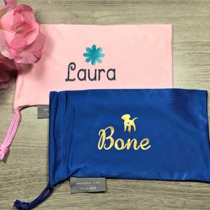 May include: Two personalized eyeglass cases. One is pink with a teal flower and the name "Laura" embroidered on it. The other is blue with a gold dog silhouette and the name "Bone" embroidered on it.