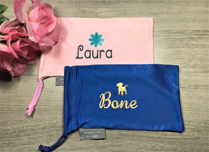 May include: Two personalized eyeglass cases. One is pink with a teal flower and the name "Laura" embroidered on it. The other is blue with a gold dog silhouette and the name "Bone" embroidered on it.
