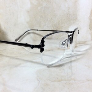 May include: A pair of semi-rimless eyeglasses with black frames and clear lenses. The glasses have a delicate design with small decorative elements on the temples. The lenses are clear, and the nose pads are visible. The glasses are set against a light-colored background.