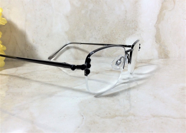May include: A pair of semi-rimless eyeglasses with black frames and clear lenses. The glasses have a delicate design with small decorative elements on the temples. The lenses are clear, and the nose pads are visible. The glasses are set against a light-colored background.